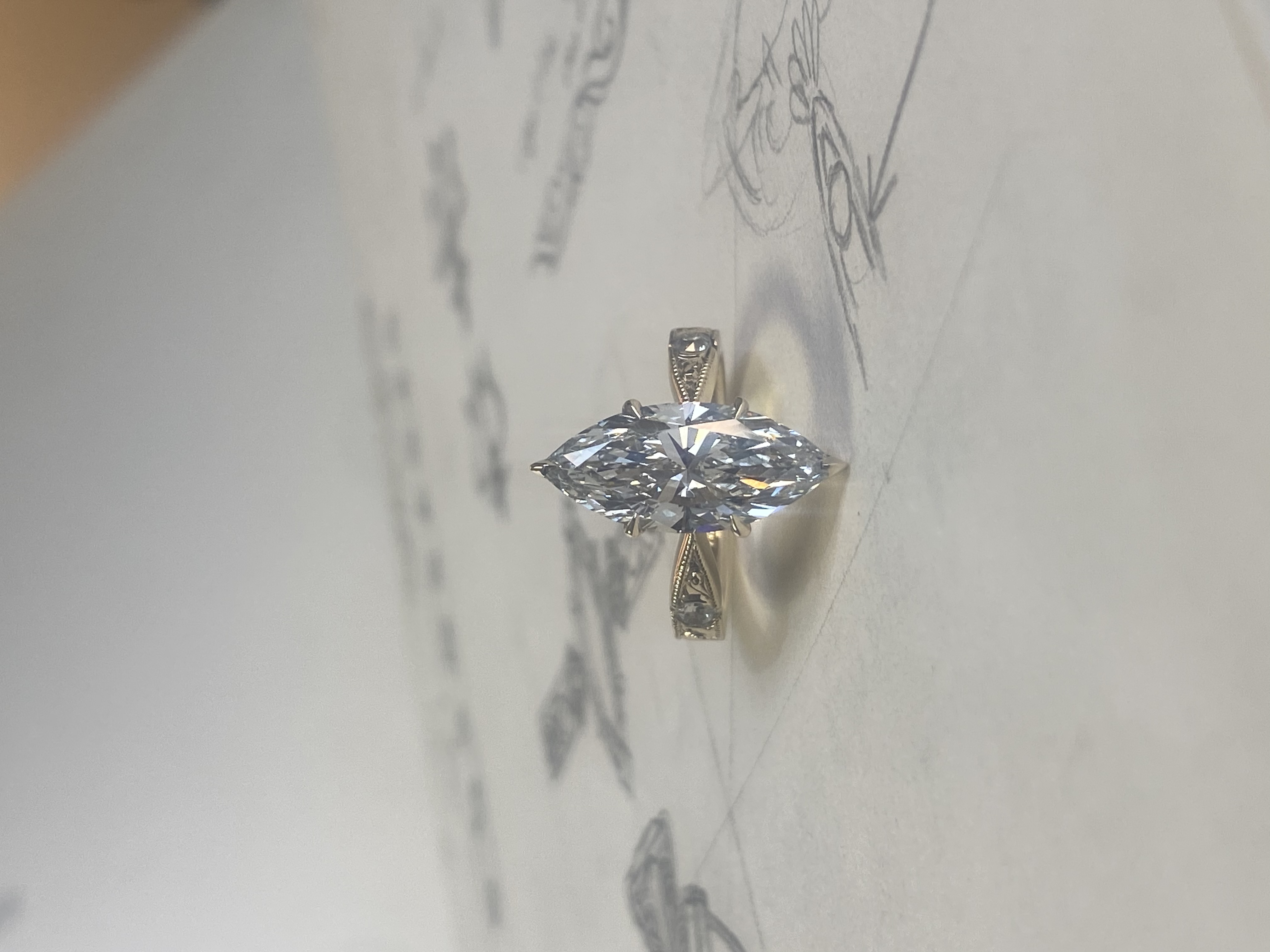 Custom marquise diamond engagement ring with aquatic shell hand-engraving, resting on original design sketches — Bear Goldsmithing Toronto